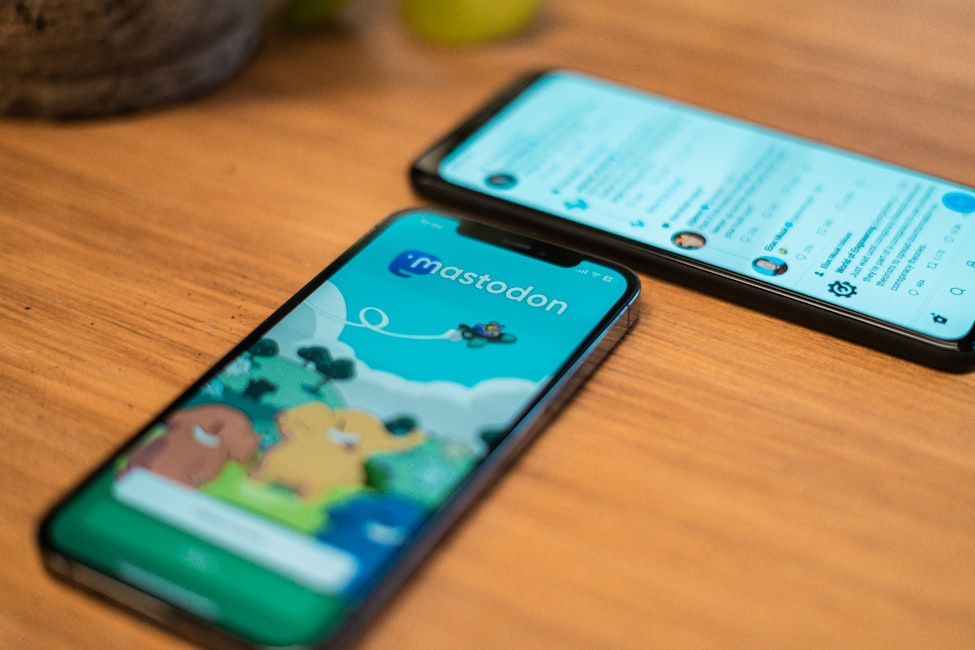 A picture of two phones laying on a wooden desk. Both showing the official Mastodon app.